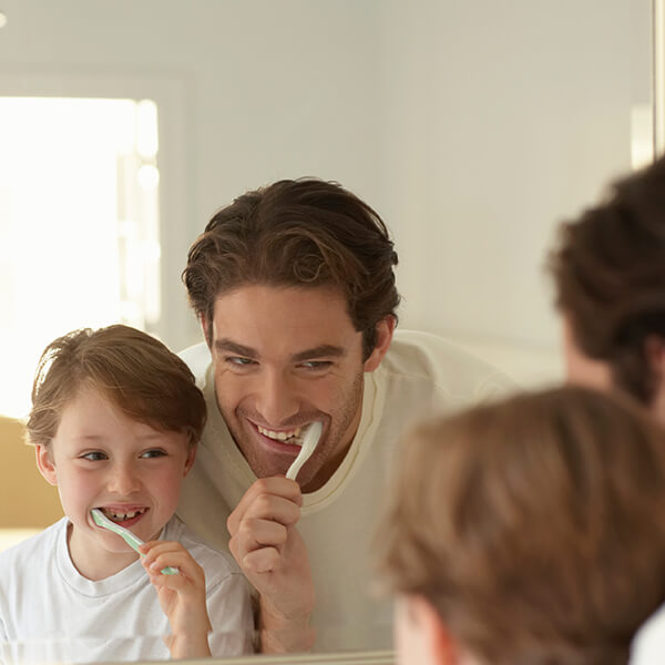 A dad brushing his teeth next to his little son while they are reflected in the mirror